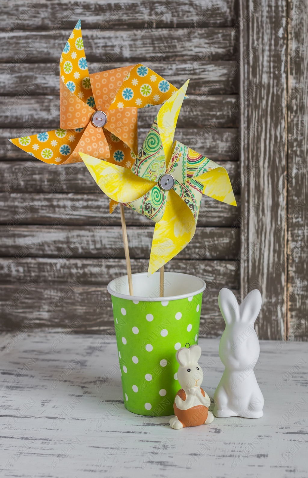 Homemade paper pinwheel, ceramic bunnies on a light rustic wood table. Easter still life in vintage 