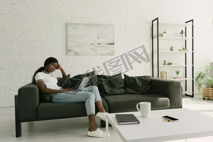 thoughtful african american freelancer sitting on sofa with laptop in spacious living room