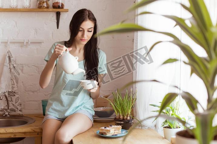 Beautiful brunette woman sitting in the kitchen, on the table, drinking tea and eating waffle, desse