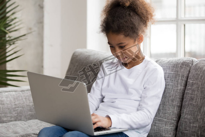 Curious preschool african mixed race girl using laptop on couch, little art black kid typing on co