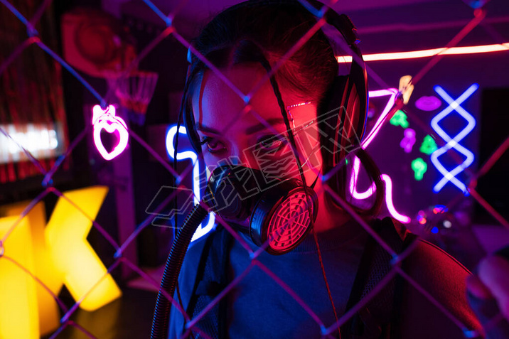 young asian woman in gas mask and wireless headphones near blurred metallic fence 