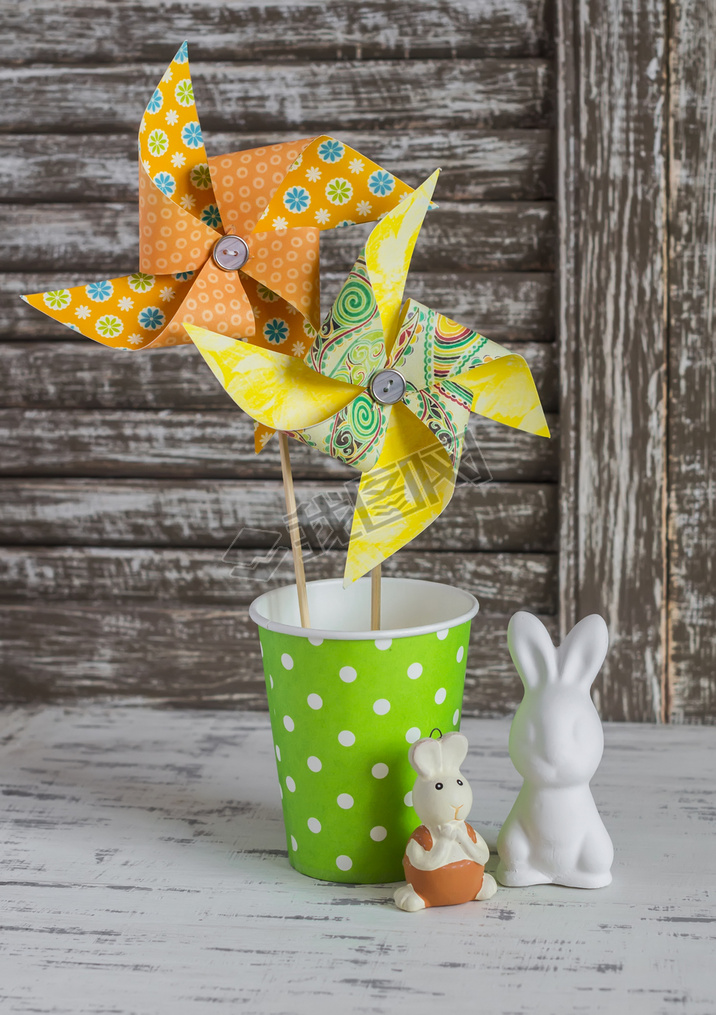 Homemade paper pinwheel, ceramic bunnies on a light rustic wood table. Easter still life in vintage 