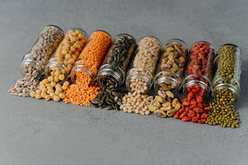 Dried seeds, beans and various grains poured out from glass bottles, spread on grey background. Heal