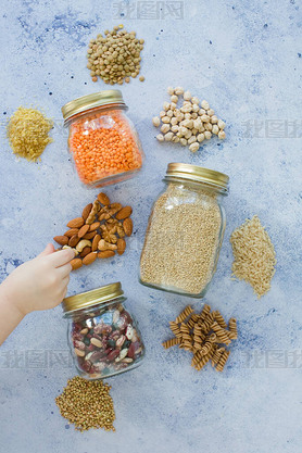 Cereal grains, seeds, beans in mason jars on a blue background. Child's hand touch takes nuts, healt