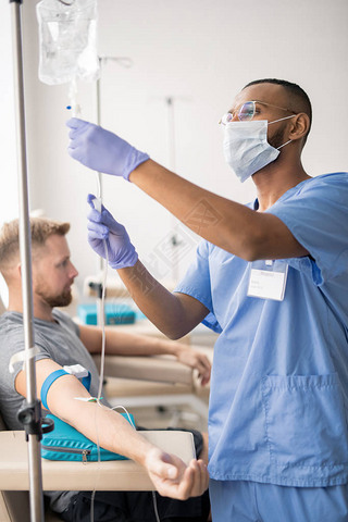 Young mixed-race doctor in blue uniform, protective mask and rubber gloves preparing dropper for one