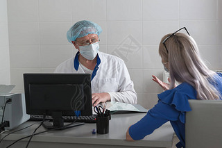 A doctor in a white coat, mask and cap in the office sits at a table with a patient and listens to h