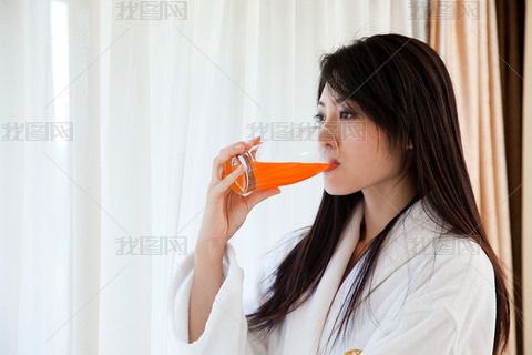 穿浴袍的年轻女人站窗边喝果汁