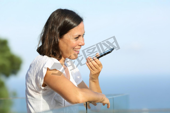 Happy adult woman using voice recognition on art phone standing in a balcony on the beach