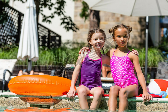 girls in swimsuits hugging while sitting at poolside near inflatable ball and ring