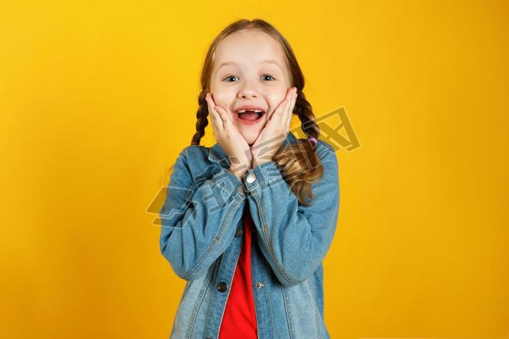 Cheerful emotional little girl in a denim shirt on a yellow background. The child is pleased with th