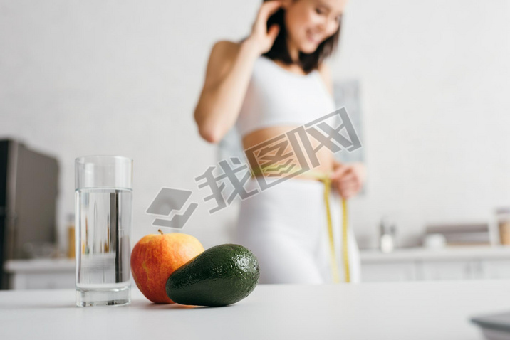 Selective focus of glass of water and fruits on table and iling sportswoman measuring waist with t