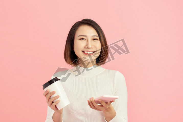 Close up portrait of young Asian woman holding coffee cup and artphone over pink background.