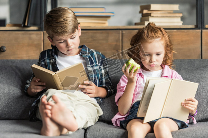 concentrated little brother and sister reading books on couch at home