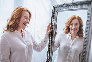 beautiful iling mature woman standing near mirror and looking at reflection