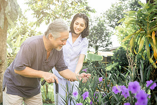 Comforting hand. Young nurse holding old man's shoulder in outdoor garden looking at flower. Senior