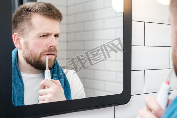 Serious man brushing his teeth with an electric oral irrigator with a jet of water, looking in the m