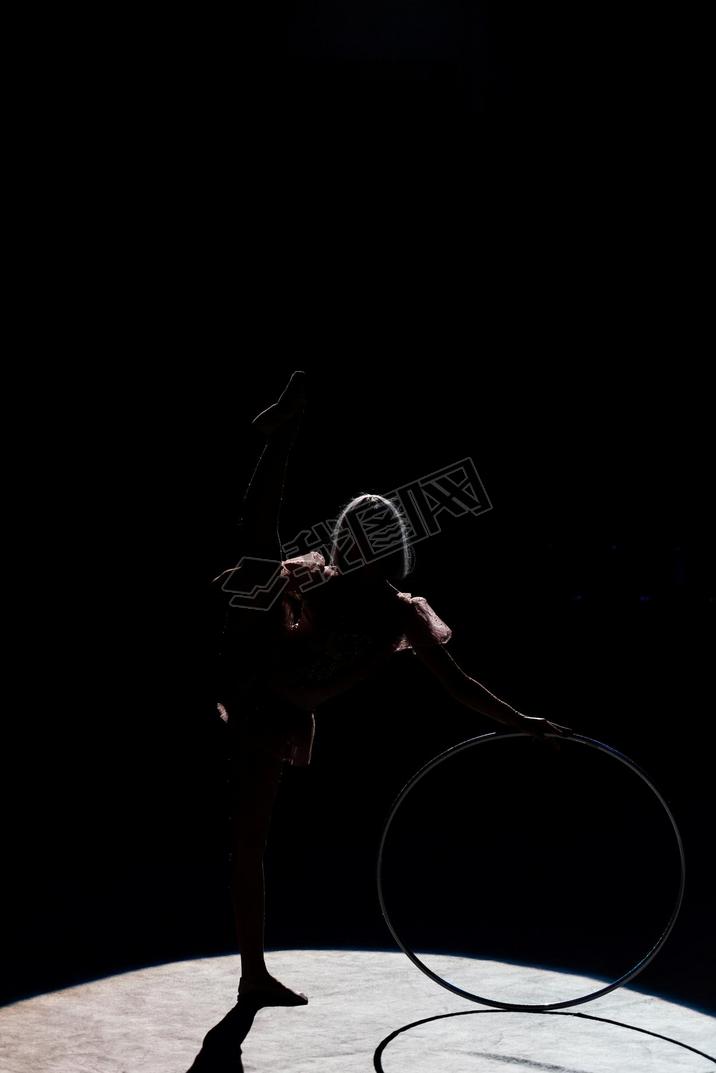 silhouette of flexible acrobat standing with hula hoop in arena of circus 