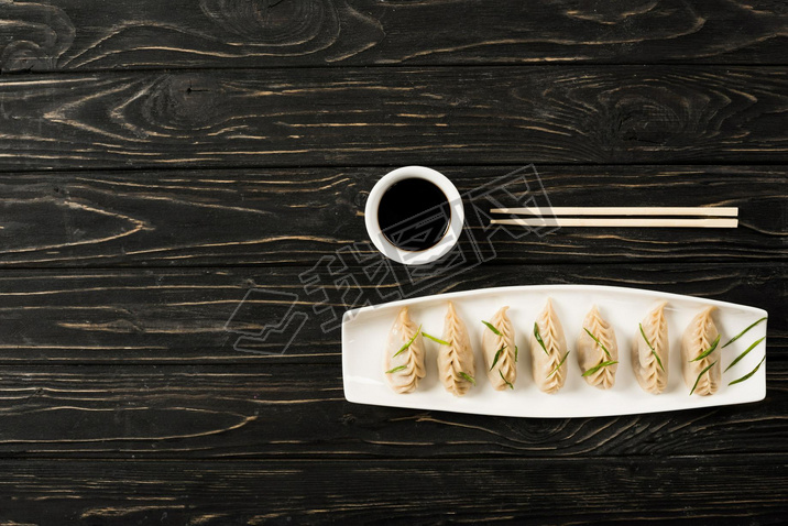 top view of delicious Chinese boiled dumplings on plate near chopsticks and soy sauce on black woode