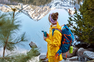 Hiking woman stops near lake in mountains, carries backback, holds thermos of hot beverage, explores