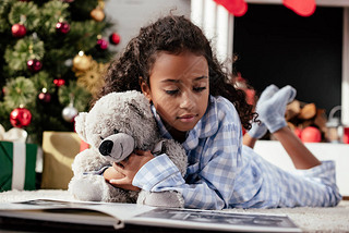 adorable african american child in pajamas with teddy bear looking at photo album on floor at home