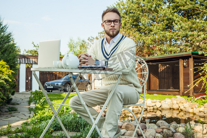 Young stylish handsome man in bright confidential clothes work at iron table with computer  teapot 