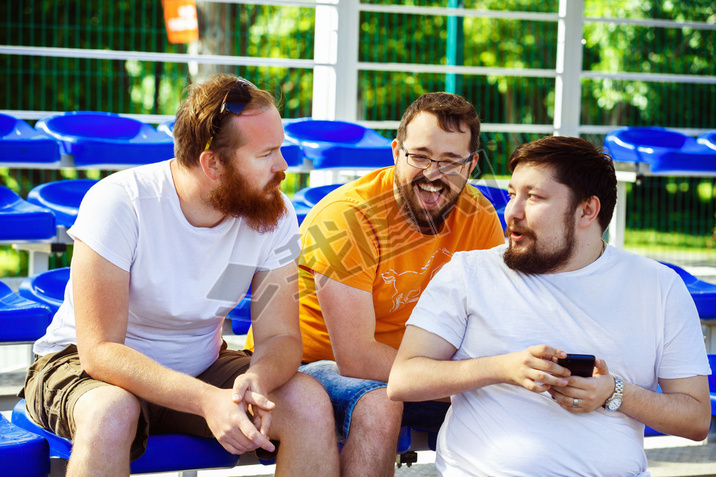 Three male friends are watching mobile phone video and laughing sitting at summer day at stadium bac