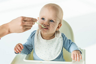 Mother with spoon feeding cute baby boy on feeding chair on white background