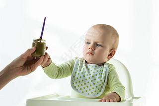 Selective focus of mother giving jar of baby nutrition to infant on feeding chair on white backgroun