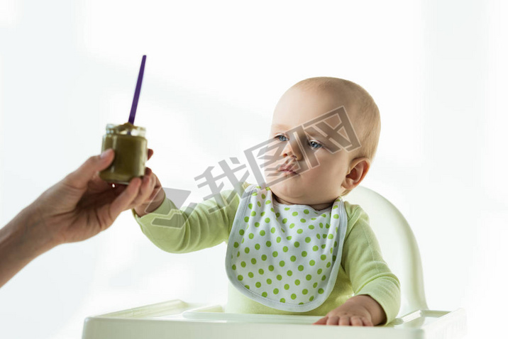 Selective focus of mother giving jar of baby nutrition to infant on feeding chair on white backgroun