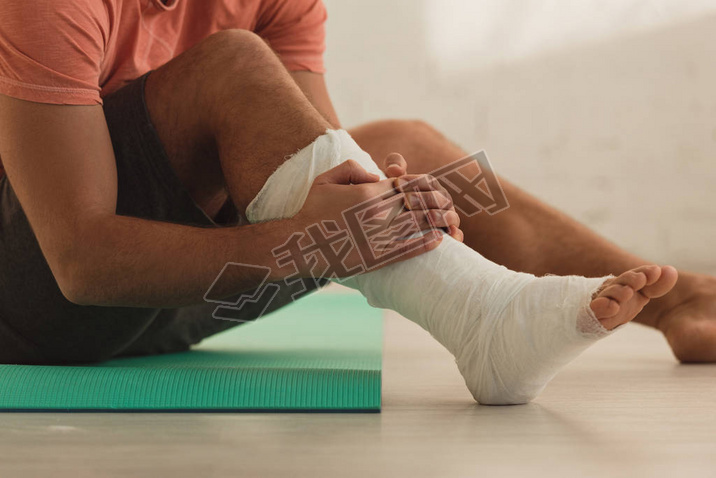 Cropped view of man holding broken leg and sitting on fitness mat on floor
