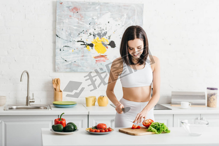 Smiling sportswoman cooking salad with ripe vegetables on kitchen table