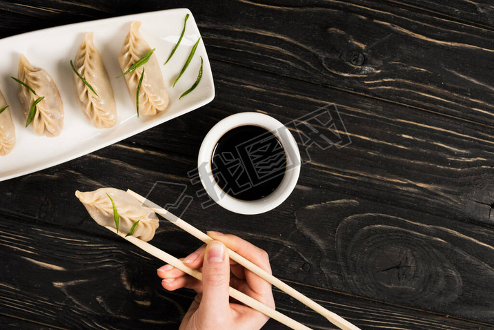 cropped view of woman eating delicious Chinese boiled dumpling with chopsticks and soy sauce at blac