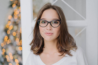 Headshot of lovely young female model in transparent glasses, has dark hair, dressed in casual cloth