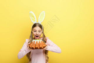 shocked kid with bunny ears holding easter cake isolated on yellow