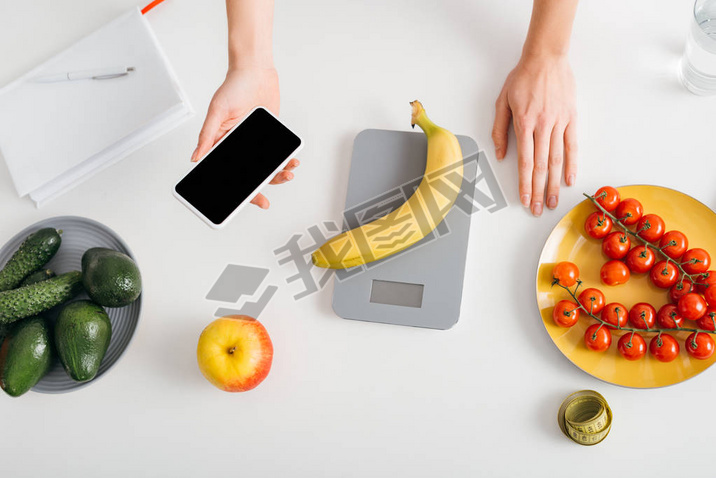 Top view of girl holding artphone while weighing banana on kitchen table, calorie counting diet