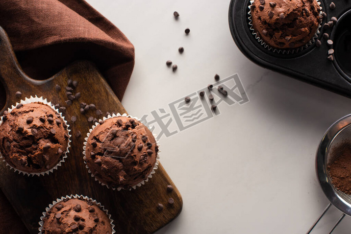 top view of fresh chocolate muffins on wooden cutting board near brown napkin and cocoa powder on ma