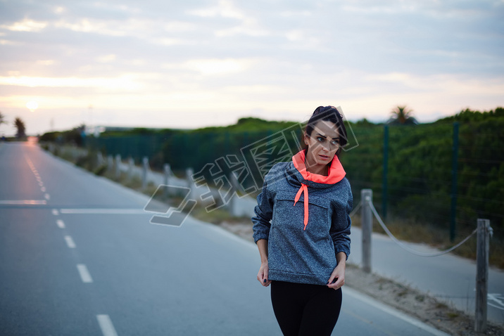 Fitness woman dressed in bi color hoodies standing on the road at sunset