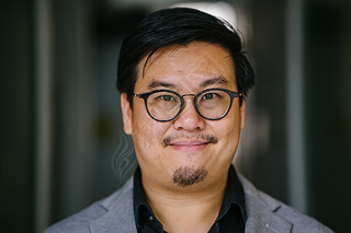 一位受过教育和中年中国亚裔 (新加坡) 男子的肖像, 穿着黑色衬衫和灰色西装。当他凝视着相机时, 他在微笑。.