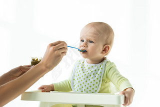 Mother with jar of vegetable baby nutrition and spoon feeding infant on feeding chair isolated on wh