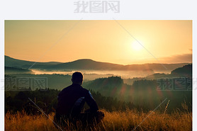 Hiker in squatting position in high grass meadow  enjoy the colorful sunrise scenery