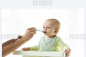 Mother with jar of vegetable baby nutrition and spoon feeding infant on feeding chair isolated on wh