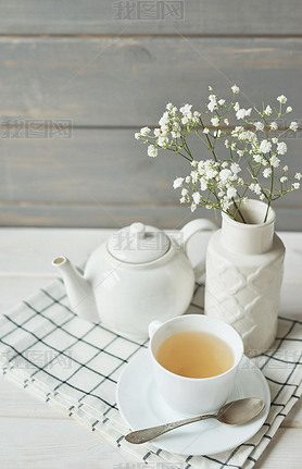 green tea in a cup next to a teapot and a vase with baby's breath on a wooden background