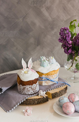 Easter Cakes on wooden decorated with rabbit ears, eggs on whith plate on foreground, lilac on backg