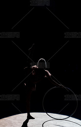 silhouette of flexible acrobat standing with hula hoop in arena of circus 