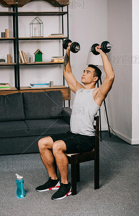 strong mixed race sportan holding hey dumbbells over head while sitting on chair