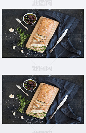 Italian ciabatta bread cut in slices on wooden chopping board with herbs, garlic and olives over dar