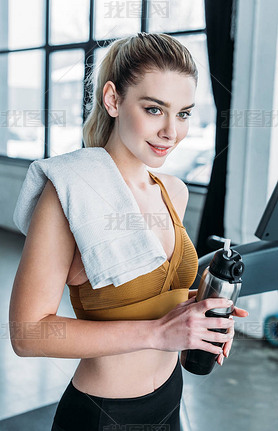 beautiful iling young sportswoman with towel on shoulder holding sports bottle in gym