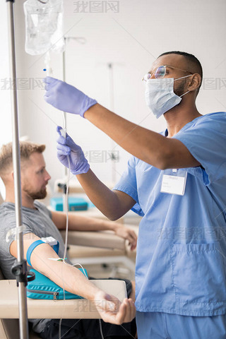 Young mixed-race doctor in blue uniform, protective mask and rubber gloves preparing dropper for one