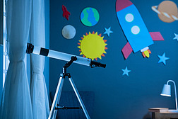 夜间在小孩房间里的三脚架上的望远镜。望远镜站在靠近窗户的儿童卧室里，墙上装饰着硬纸板火箭和太阳系。带有行星、恒星和月球装饰的带有复制空间的墙壁的儿童房的内部.