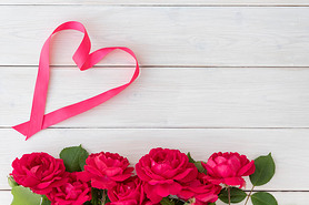 Top view of red roses on bottom and heart shape made from ribbon on white wooden background, greetin